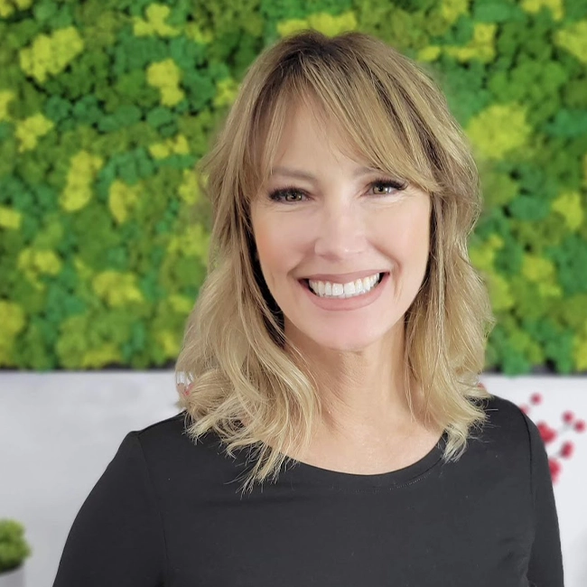 A headshot of Mindy smiling in front of the EDEN Day Spa green moss background board